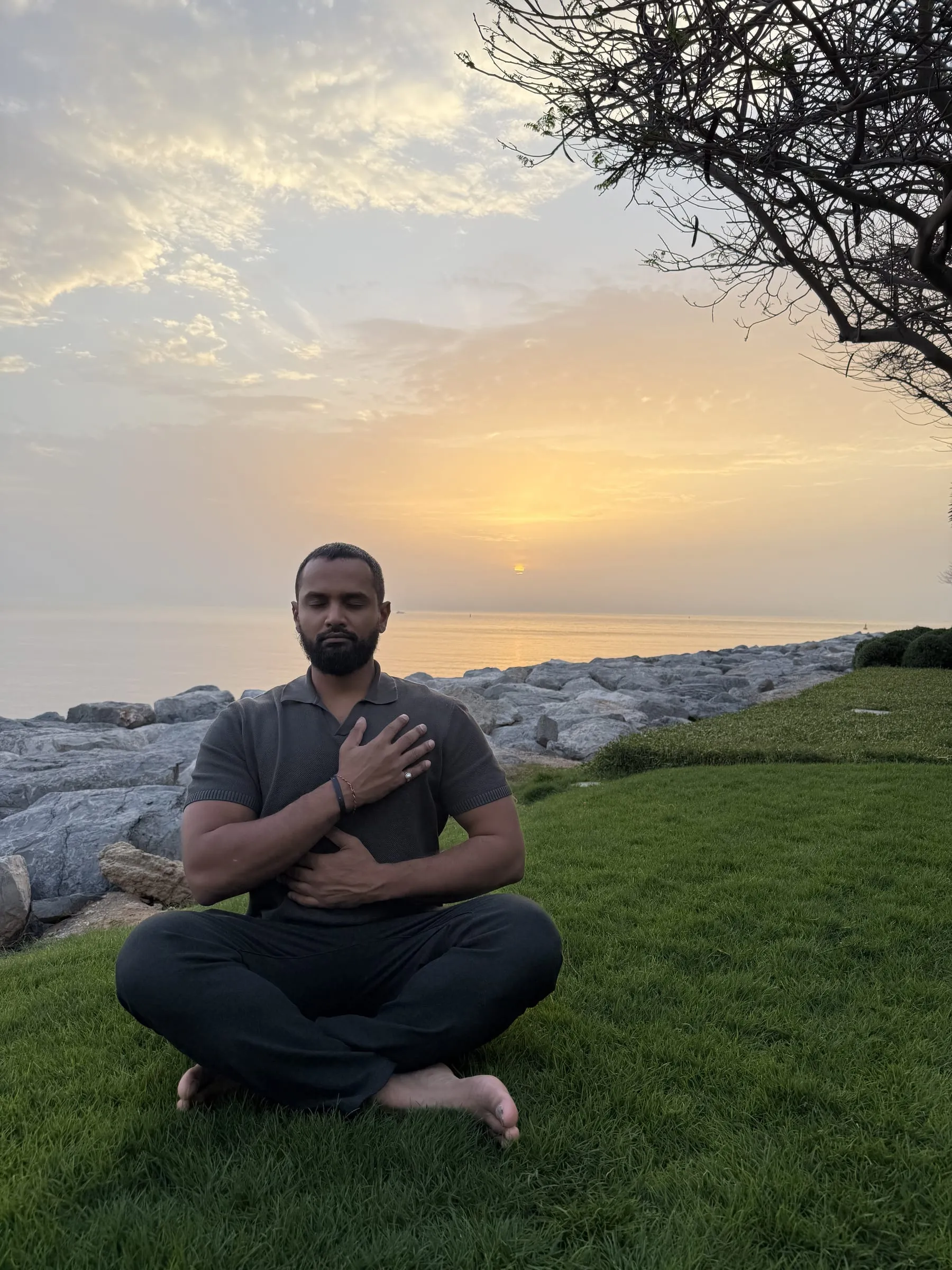 Pranav Shenoy sitting cross-legged on grass by the sea at sunrise, one hand resting on his chest.