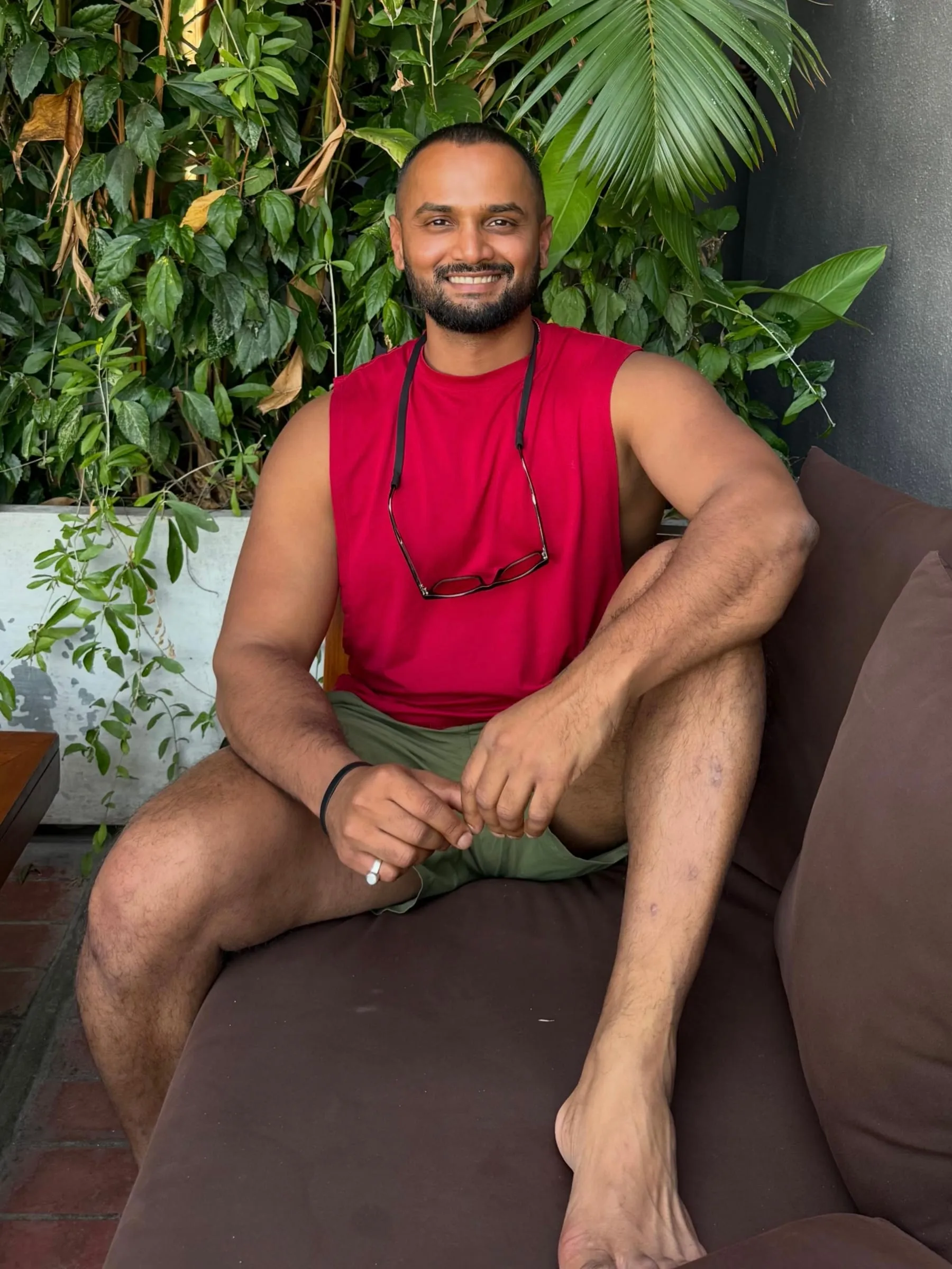 Pranav Shenoy sitting on a couch outdoors, smiling, with tropical planting behind him.